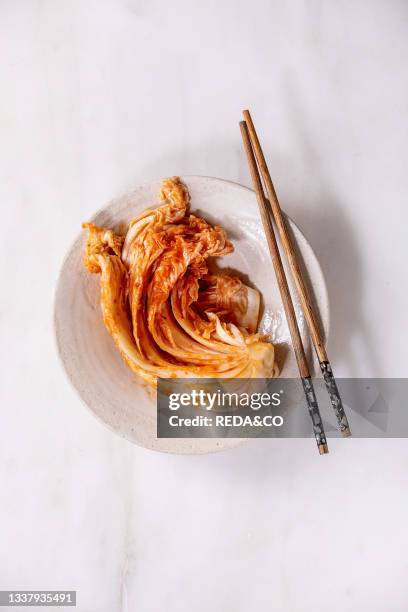 Homemade korean traditional fermented appetizer kimchi cabbage served in ceramic plate with chopsticks over white marble background. Flat lay. Copy...