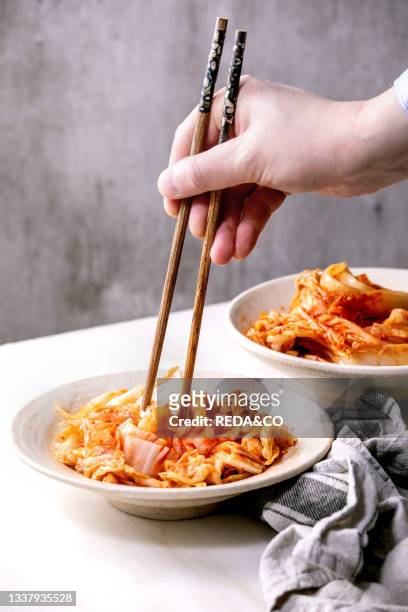 Homemade korean traditional fermented appetizer kimchi cabbage served in ceramic plate over white marble background. Chopsticks in mans hands.