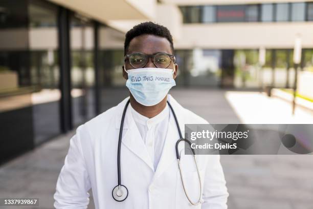 porträt eines jungen männlichen arztes, der während der covid-19-pandemie vor dem krankenhaus steht und eine schutzmaske trägt. - protestor mask stock-fotos und bilder