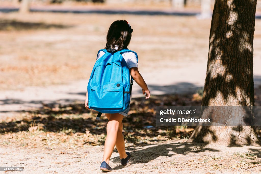 A black-haired girl with a blue backpack, turned her back, walks through a park in the direction of school. Concept of education and back to school.