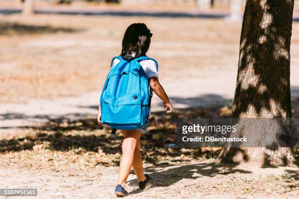 a black-haired girl with a blue backpack, turned her back, walks through a park in the direction of school. concept of education and back to school. - satchel bag stock pictures, royalty-free photos & images