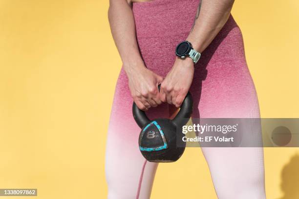 unrecognizable strong woman in pink fitness sportswear lifting weight kettlebell over yellow background - kettle bells stockfoto's en -beelden