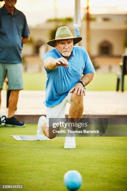 Wide shot of senior man throwing bowl during lawn bowling match on summer evening