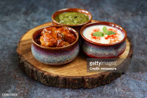 image of three bowls containing dips of raita (yoghurt based sauce), mint coriander chutney and green mango pickle, on wooden chopping board, mottled grey background, indian cuisine, focus on foreground - 美味食品 個照片及圖片檔