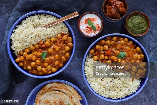 image of two blue plates containing homemade punjabi chole (chickpea curry) meals served with white rice and lachha paratha (flatbread), bowls of green mango chutney, raita, mint and coriander dip, fork, mottled blue background, elevated view - curry powder stock pictures, royalty-free photos & images