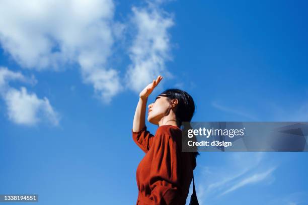 low angle portrait of young asian woman shielding eyes with her hand, standing against beautiful clear blue sky and sunlight. freedom in nature. connection with nature - health shield stock pictures, royalty-free photos & images