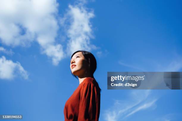 low angle portrait of confident young asian woman standing against beautiful clear blue sky, looking up to sky enjoying sunlight. freedom in nature. connection with nature - project greenlight stock pictures, royalty-free photos & images