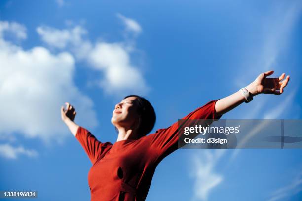 low angle portrait of beautiful young asian woman with her eyes closed stretching arms, setting herself free and feeling relieved. enjoying fresh air and sunlight with head up against beautiful clear blue sky. freedom in nature. connection with nature - fresh air breathing stockfoto's en -beelden