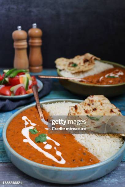 close-up image of blue bowls containing homemade dal makhani (black lentils and red kidney bean curry) meals, served with white rice and naan flatbread, metal spoons, side salad, salt and pepper shakers, blue wood grain surface, focus on foreground - curry leaves stockfoto's en -beelden