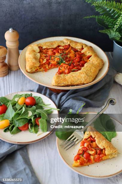 bild von einem stück hausgemachtem herzhaftem galette-rezept auf weißem teller mit gabel und beilagensalat, neben rustikalem kuchen auf kuchenständer, pesto, kirschtomatenhälften, zerbröckeltem feta-käse und rosmarinblättern in blätterteigkruste, g - biscotte stock-fotos und bilder