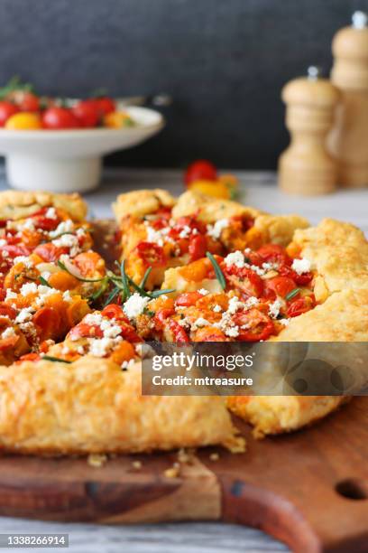 imagen de galette salada casera en rodajas en rodajas en tabla de cortar circular de madera junto a tomates de tazón, pesto, mitades de tomate cherry, queso feta desmenuzado, hojas de romero en corteza de hojaldre, superficie de grano de madera gris, enfo - galette fotografías e imágenes de stock