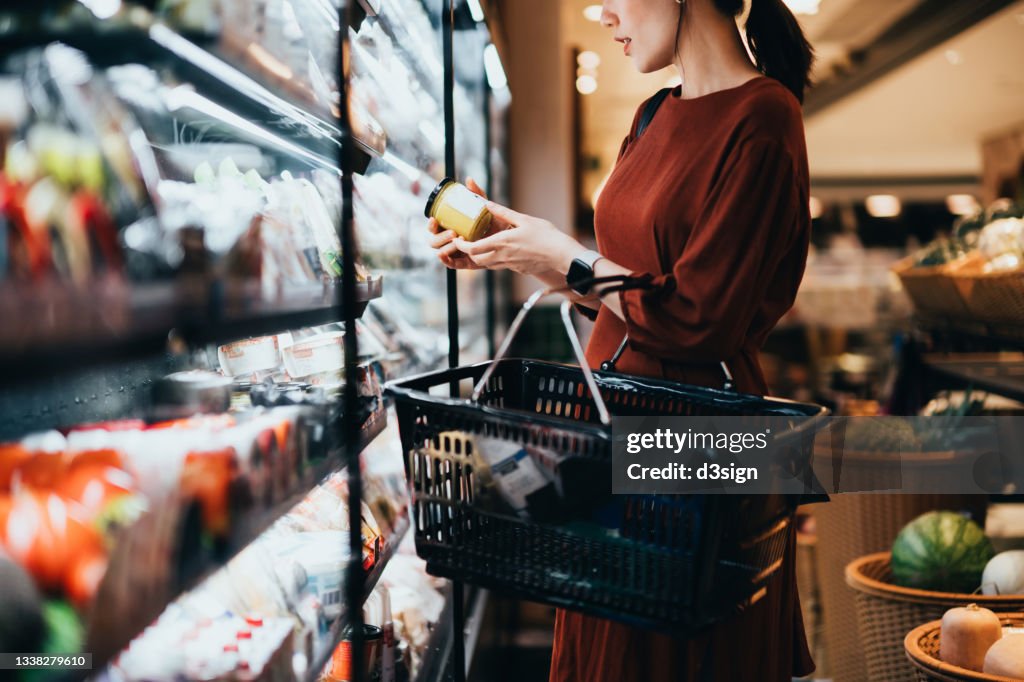Cropped shot of young Asian woman carrying a shopping basket, standing along the dairy aisle, reading the nutrition label on the bottle of a fresh organic healthy yoghurt. Making healthier food choices