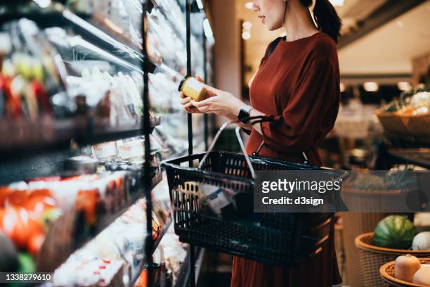 cropped shot of young asian woman carrying a shopping basket, standing along the dairy aisle, reading the nutrition label on the bottle of a fresh organic healthy yoghurt. making healthier food choices - speisen und getränke stock-fotos und bilder