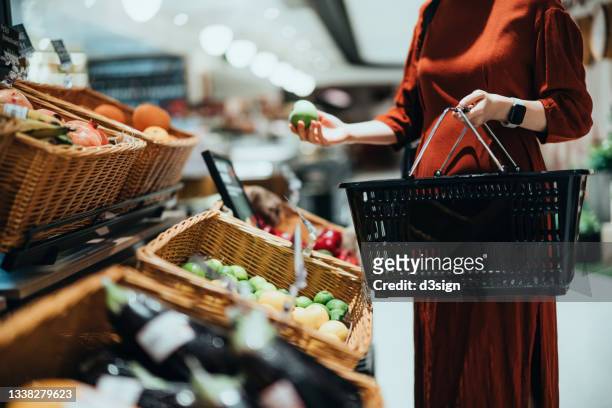 cropped shot of young asian woman carrying a shopping basket, grocery shopping for fresh organic fruits and vegetables in supermarket. green living. making healthier food choices - woman supermarket stockfoto's en -beelden