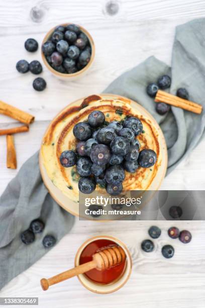 image stack of american-style blueberry pancakes with fresh berries, bowls of berries and honey with wooden honey dipper, grey muslin, surrounded by fresh blueberries, marble effect background, elevated view - blueberry pancakes stock pictures, royalty-free photos & images