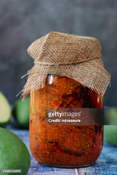 image glass jar of green mango chutney recipe with hessian lid, pickle with chopped raw mango, seeds, spices and mineral condiments, blue wood grain surface, grey background, focus on foreground - sliced pickles stockfoto's en -beelden