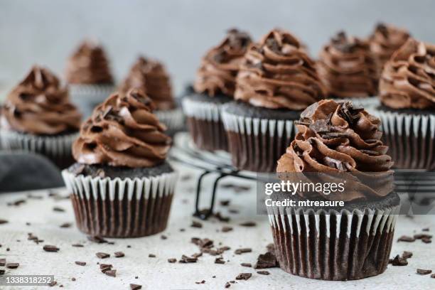 close-up image of batch of homemade, chocolate cupcakes in paper cake cases on circular, black, metal wire cooling rack, chocolate butter icing piped swirls topped with chocolate pieces, grey muslin, white background, focus on foreground - cupcake stock pictures, royalty-free photos & images