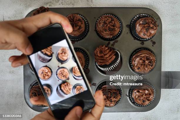 image of unrecognisable person using smartphone  to photograph muffin cake tin containing homemade, chocolate cupcakes in paper cake case, chocolate butter icing piped swirl topped with chocolate pieces, cakes lying on side, elevated view - cake case stock pictures, royalty-free photos & images