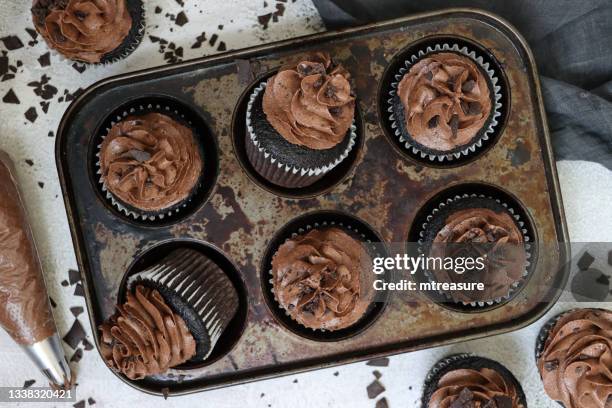 image of muffin baking tray with homemade, chocolate cupcakes in paper cake cases, chocolate butter icing piped swirl topped with chocolate pieces, white background, cake tin with baked on dirt surrounded by cakes, piping bag, grey muslin, elevated view - baking dish stock pictures, royalty-free photos & images