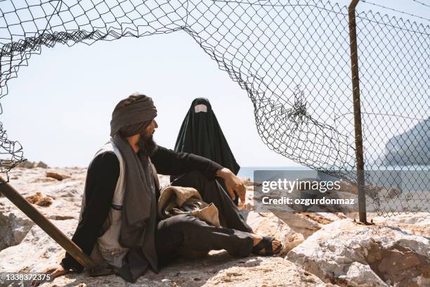 refugee man and woman in burka standing behind a fence - evacuatiekamp stockfoto's en -beelden