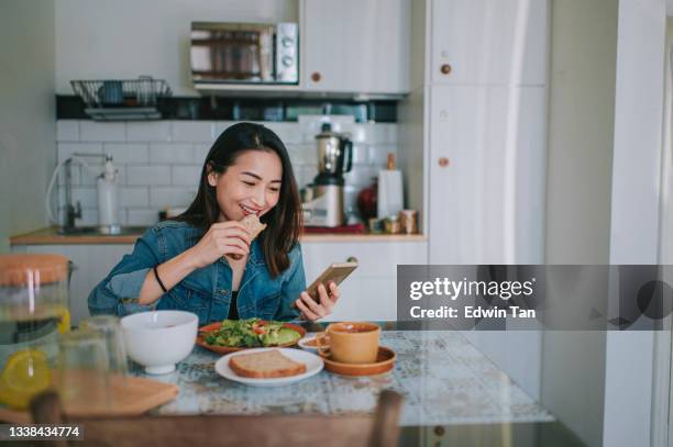 asian chinese beautiful woman reading on her smart phone and enjoying her breakfast at home buttering wholegrain bread peanut butter with coffee - asian females on a phone imagens e fotografias de stock