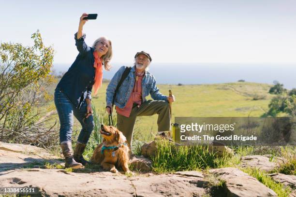 senior couple taking selfie with dog during hike on sunny day - active seniors foto e immagini stock