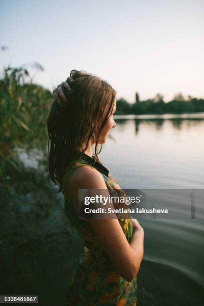 a young teenage girl with wet hair and a wet colorful dress stands on the shore of the lake against a background of tall grass and water in the sunset light and poses looking into the distance with her hands raised to her chest. low key photo. side view. - girls in wet dresses stock-fotos und bilder