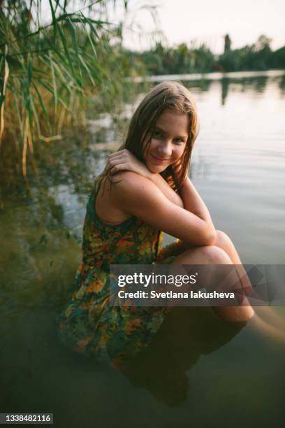 a young teenage girl with wet hair and a wet colorful dress sits in the water by the lake against a background of tall grass and water in the sunset light and poses looking at the camera, smiling with her hands raised to her chest.low key photo.side view. - girls in wet dresses stock-fotos und bilder