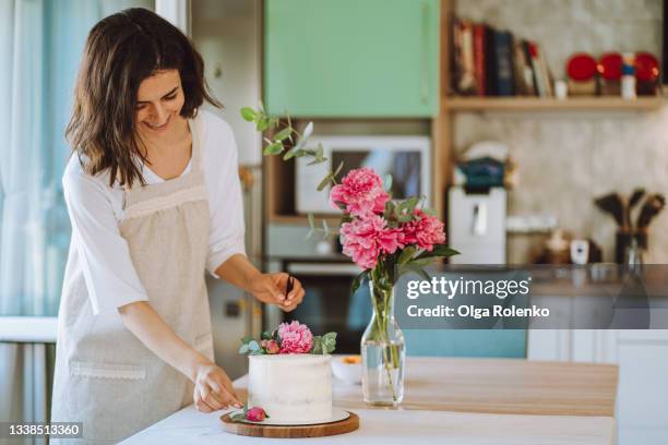 dark-haired smiling woman wearing apron decorate сake with fresh peonies and leaves in the kitchen - decorating a cake stock pictures, royalty-free photos & images
