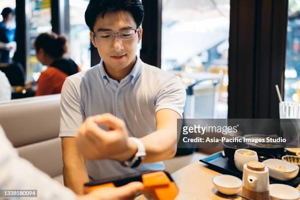young asian man enjoying japanese food and paying with smart watch in restaurant. - asian and indian ethnicities smartwatch phone stock pictures, royalty-free photos & images