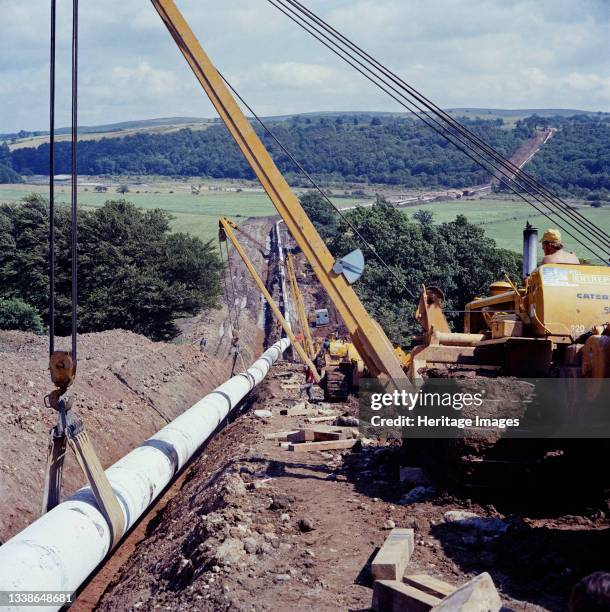 View along the route of the Brampton pipeline, showing pipelaying equipment in the foreground lowering the pipeline into a trench. In 1975, Laing...