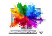 modern black silver pc monitor with mouse and keyboard colorful rainbow holi powder cloud explosion through screen isolated white background. computer multimedia abstract art streaming concept.