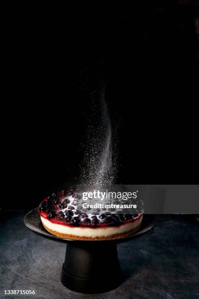 image of icing sugar being sifted onto blackcurrant cheesecake with buttery biscuit base on black cake stand, black background, white powder falling and floating mid-air, focus on foreground, copy space - cheesecake white stockfoto's en -beelden