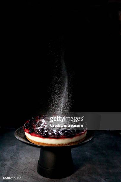 image of icing sugar being sifted onto blackcurrant cheesecake with buttery biscuit base on black cake stand, black background, white powder falling and floating mid-air, focus on foreground, copy space - black and white food 個照片及圖片檔