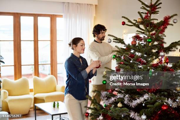 mid adult couple decorating christmas tree at home - decorare l'albero di natale foto e immagini stock