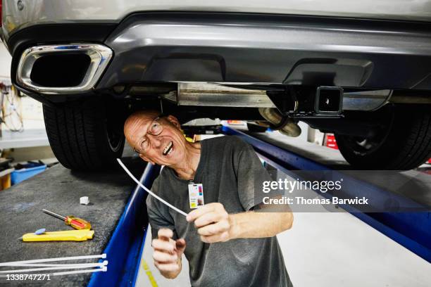 medium wide shot of laughing senior man working under car on auto lift in garage - only senior men stock pictures, royalty-free photos & images