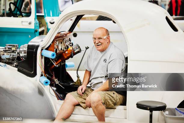 Wide shot portrait of smiling senior man sitting in auto restoration project in community garage
