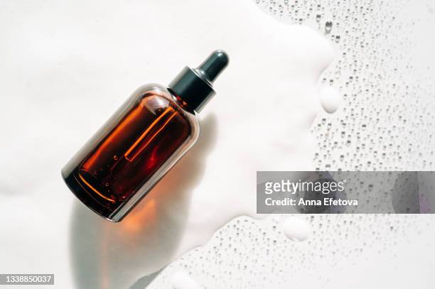 amber glass bottle with face serum on white foam among many drops. polyglutamic acid is a new hyaluronic acid. copy space for your design. flat lay style and close-up - suntan lotion stock-fotos und bilder