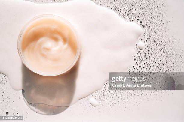 glass jar with beige cream on white foam among many drops. concept of body care and beauty. copy space for your design. flat lay style - cream dairy product stockfoto's en -beelden