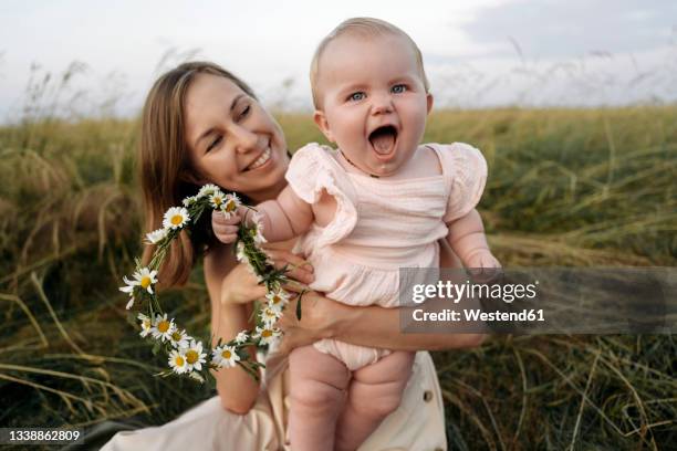 smiling mother holding cheerful baby girl with flower tiara in field - diadem imagens e fotografias de stock