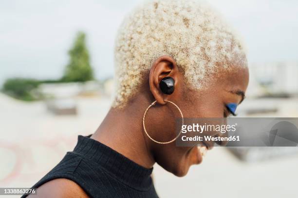 smiling young woman with wireless in-ear headphones at park - black woman short hair stock pictures, royalty-free photos & images
