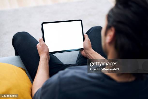 young man holding digital tablet while sitting on sofa - ipad halten stock-fotos und bilder