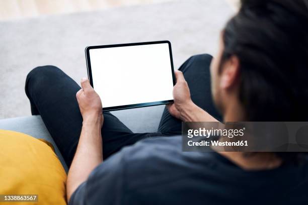 young man holding digital tablet while sitting on sofa - tablet digital imagens e fotografias de stock