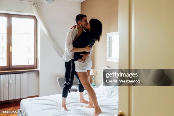 affectionate queer women kissing while standing on bed at home - gay couple in love stock pictures, royalty-free photos & images