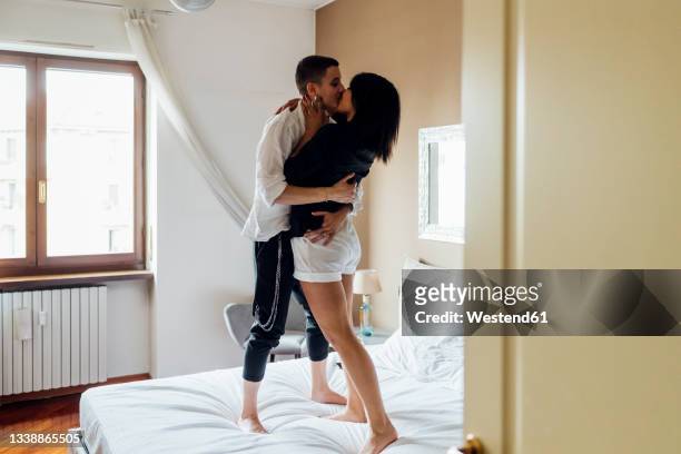 affectionate queer women kissing while standing on bed at home - couple and kiss and bedroom - fotografias e filmes do acervo