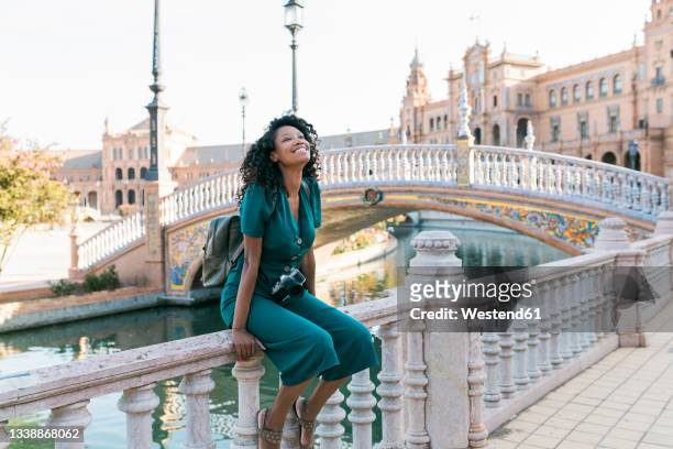 happy afro woman sitting on railing at plaza de espana, seville, spain - seville stock-fotos und bilder