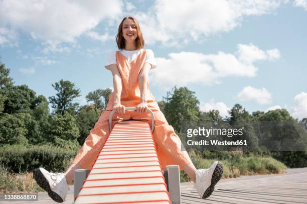 happy woman playing while sitting on seesaw at park - see saw fotografías e imágenes de stock