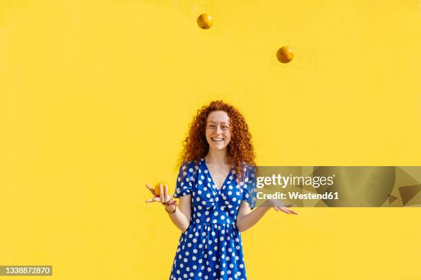 carefree woman juggling orange fruits in front of yellow wall - juggling stockfoto's en -beelden