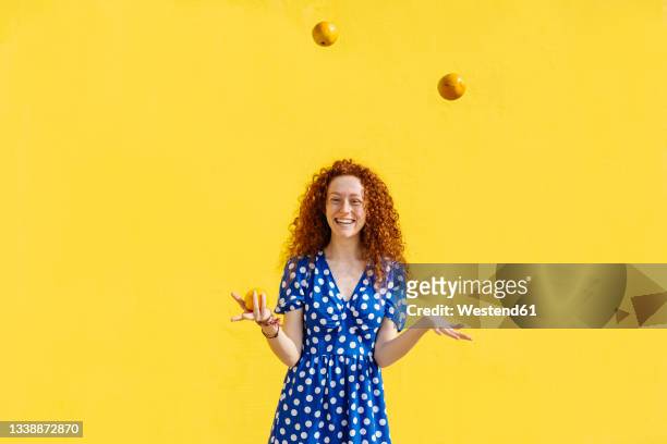 carefree woman juggling orange fruits in front of yellow wall - orange color - fotografias e filmes do acervo