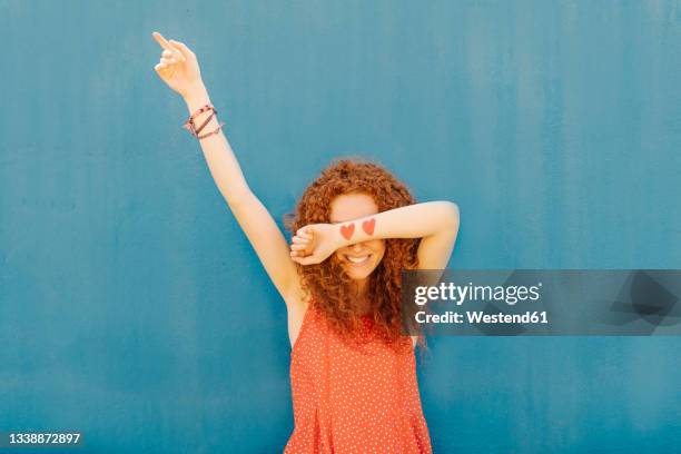 smiling woman covering eyes while standing in front of blue wall - augen zuhalten stock-fotos und bilder